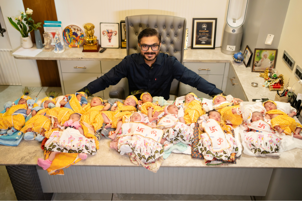 Dr. Mohil Patel seated at his clinic desk, smiling with 14 newborn babies—nine boys and five girls—dressed in festive attire, representing deliveries on Janmashtami 2025 at Ankur Maternity Home in Ahmedabad.