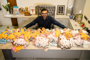 Dr. Mohil Patel seated at his clinic desk, smiling with 14 newborn babies—nine boys and five girls—dressed in festive attire, representing deliveries on Janmashtami 2025 at Ankur Maternity Home in Ahmedabad.